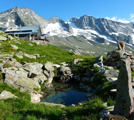 Mountain cabin on green grass with snowy peaks in the background, blue sky, small stream in front.