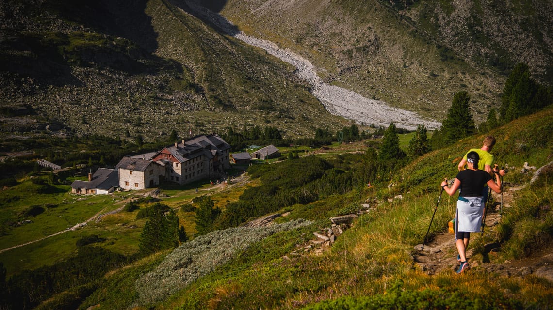 Wanderer steigen auf einem sonnigen Bergpfad zur Berliner Hütte ab. Die historische Berghütte liegt eingebettet in grüne Almwiesen und steinige Hänge im Zillertal. Der Weg ist gut sichtbar, umgeben von alpiner Naturkulisse.