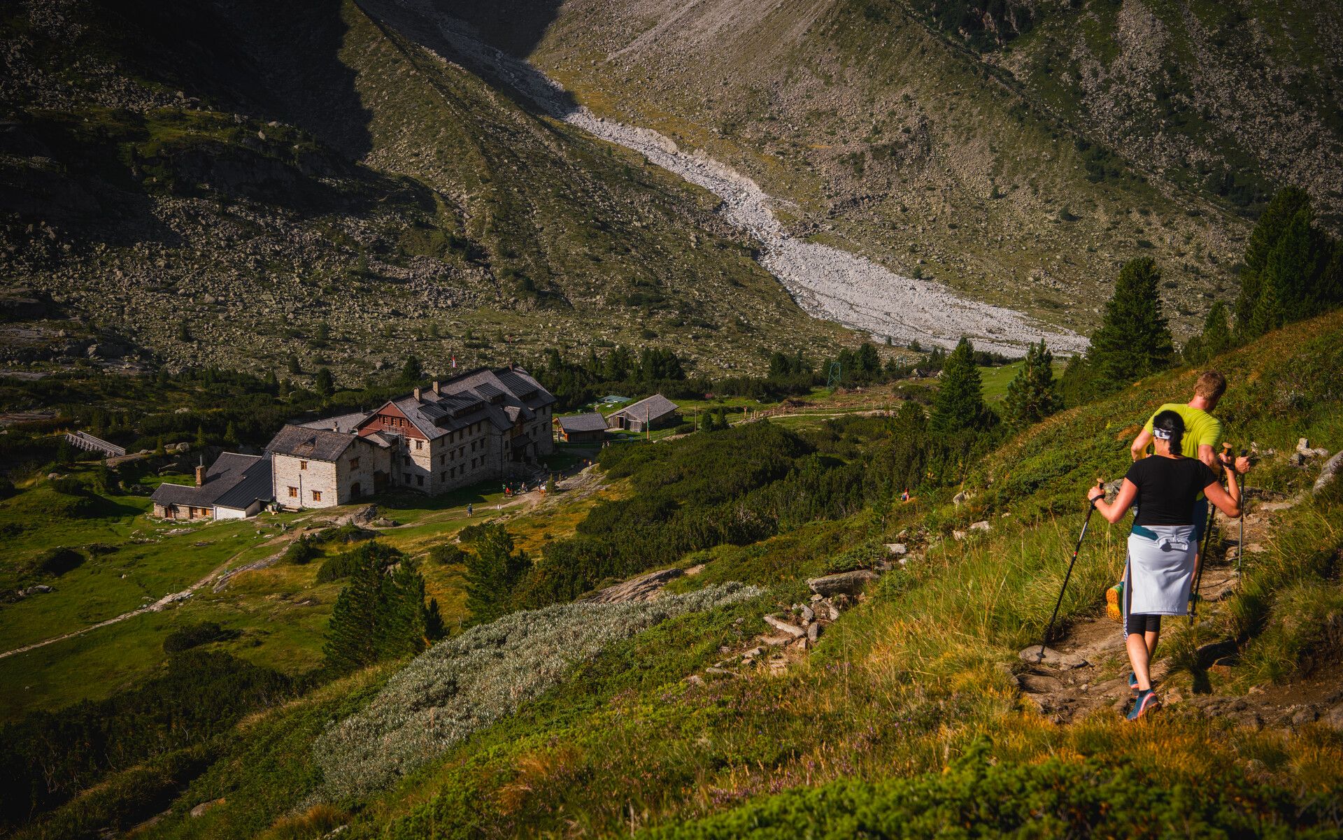 Hikers descend a sunny mountain trail toward the Berliner Hütte. The historic alpine lodge nestles in green meadows and rocky slopes in the Zillertal. The trail is clearly visible, surrounded by a pristine alpine landscape.