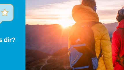 Zwei Wanderer bei Sonnenuntergang mit Rucksäcken in den Bergen, die sich unterhalten.