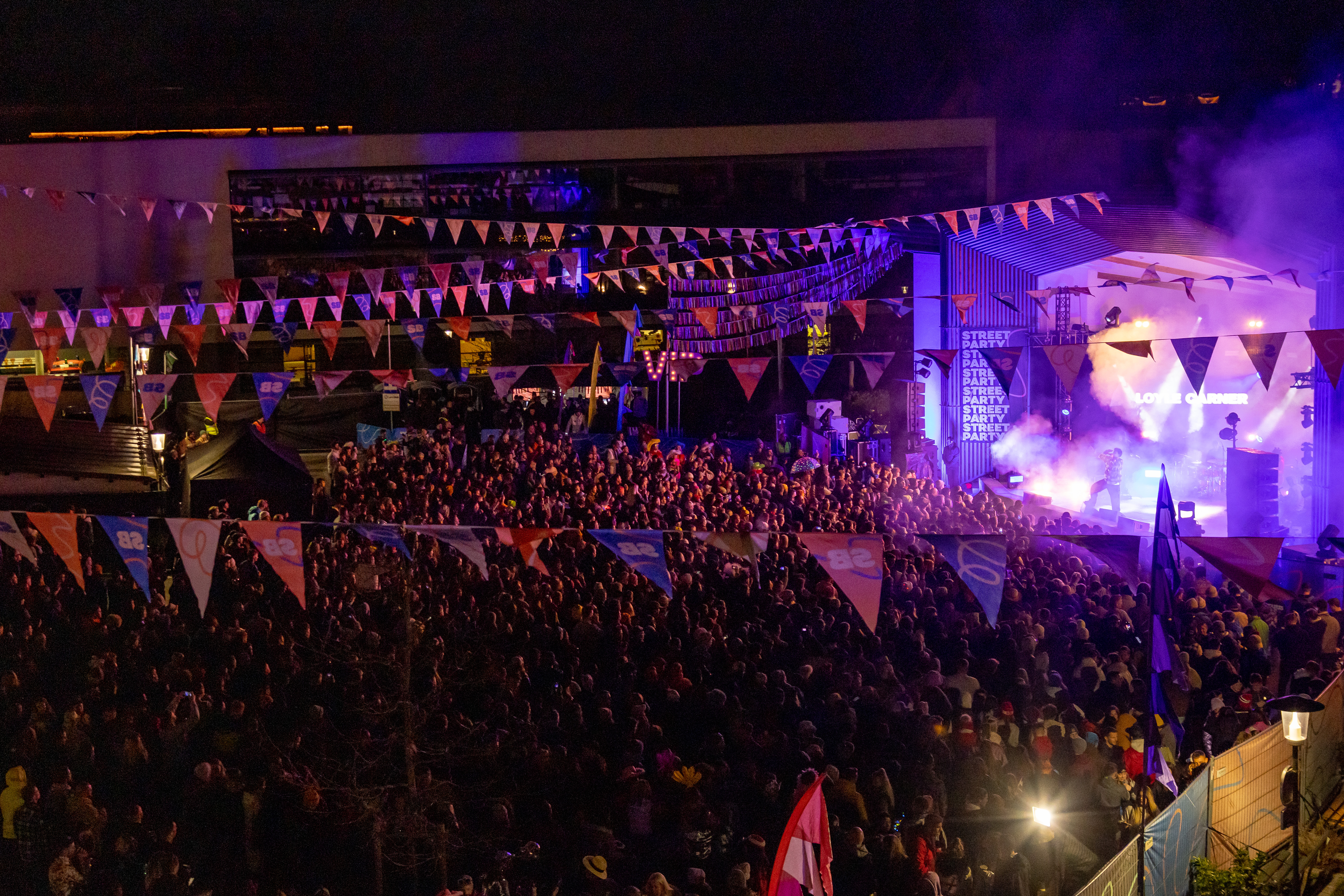 People in a large square in the evening in front of an illuminated stage; the square is decorated with flags.
