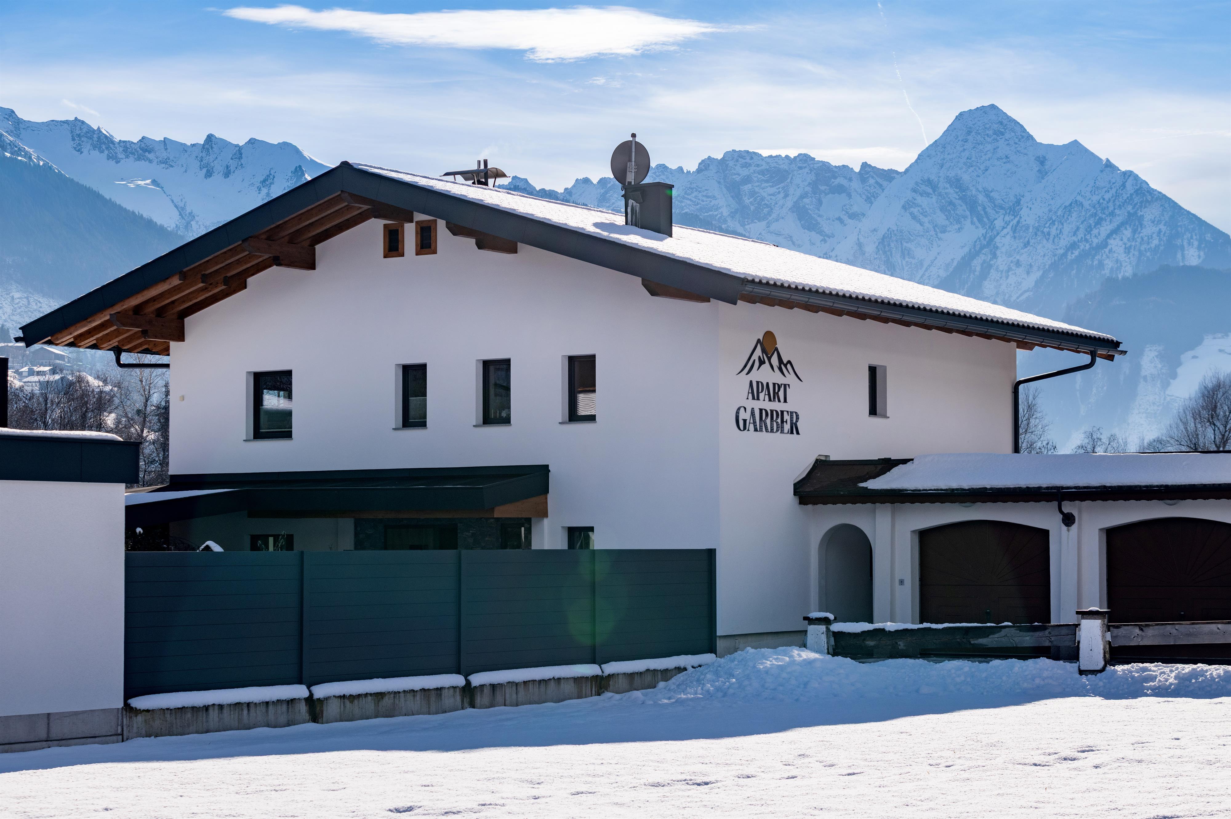 Modern house with snow-covered roof against mountain backdrop in winter, with 'Apart Garber' logo.