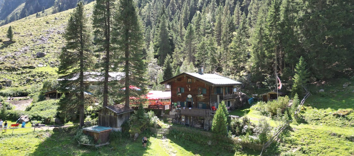 A wooden house in a lush, mountainous landscape with trees and a small garden, mountains in the background.