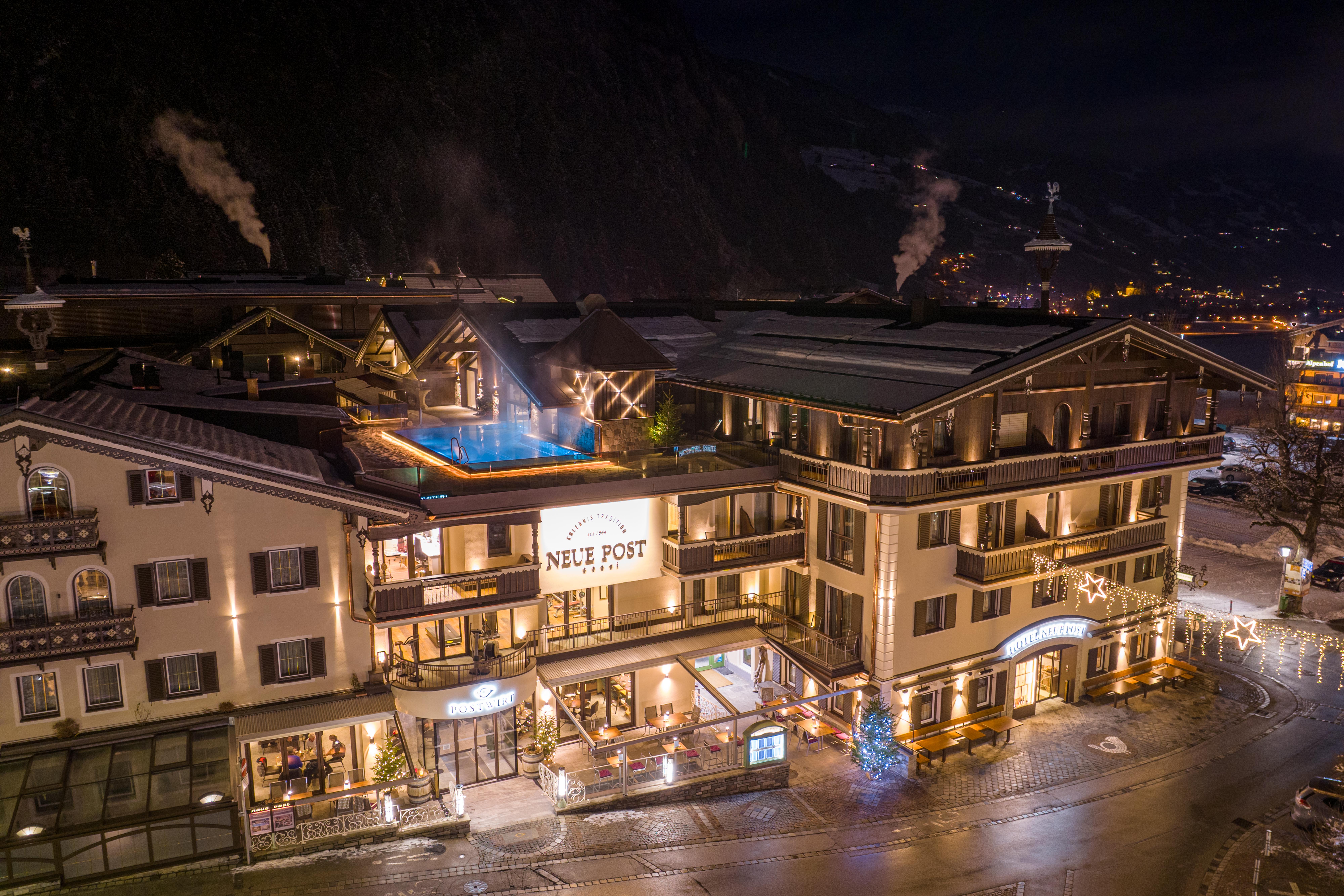 The image shows Hotel Neue Post at night from an aerial view. The illuminated hotel shines in the darkness, and the rooftop pool is clearly visible, giving the building a modern and inviting character
