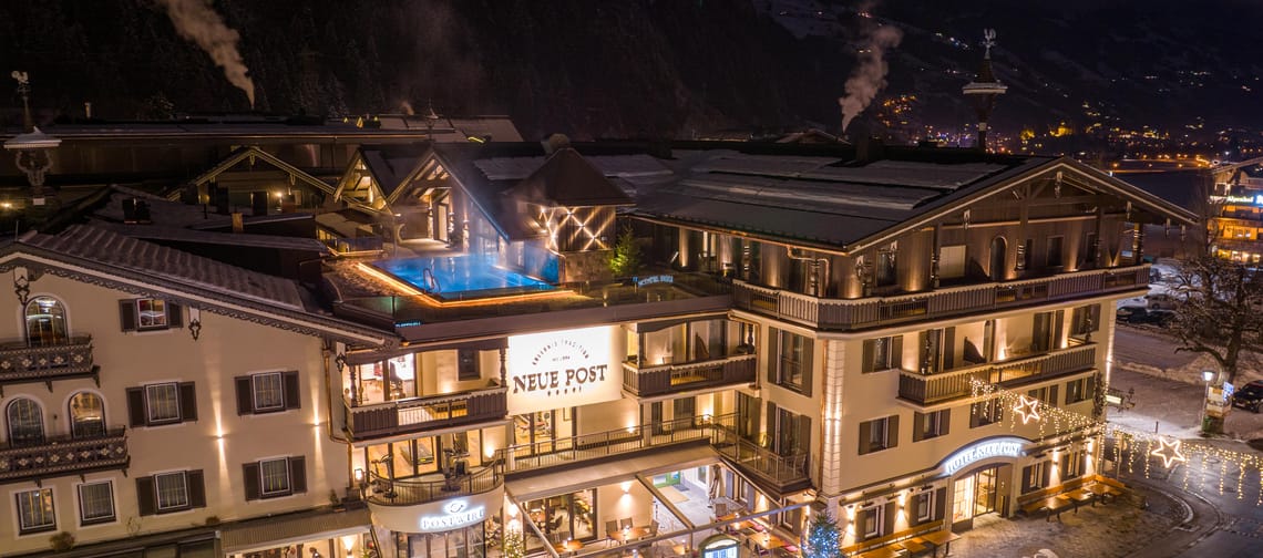 The image shows Hotel Neue Post at night from an aerial view. The illuminated hotel shines in the darkness, and the rooftop pool is clearly visible, giving the building a modern and inviting character