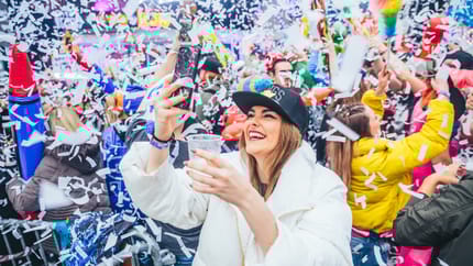 The image shows a woman at a party in the foreground wearing a white jacket, with a celebrating crowd and colorful streamers visible in the background.
