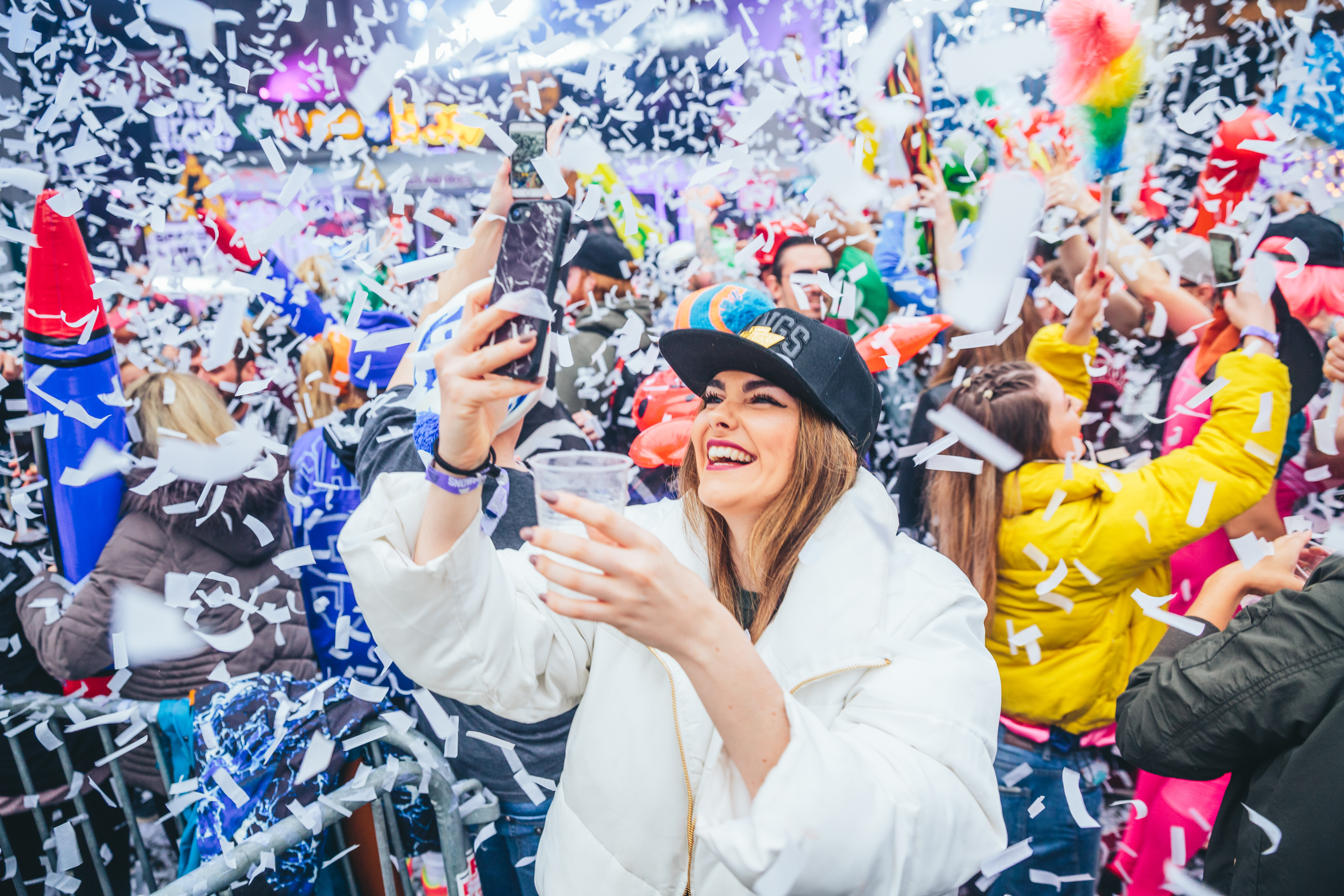 Das Bild zeigt eine Frau auf einer Party im Vordergrund mit weißer Jacke, im Hintergrund ist ein feierndes Publikum mit bunten Luftschlangen zu sehen.