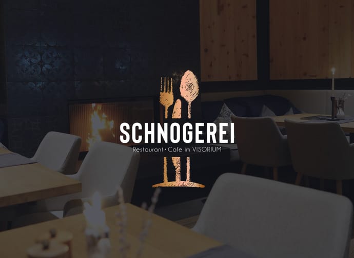 Das Bild zeigt ein gemütliches Restaurant mit moderner, stilvoller Einrichtung. Im Hintergrund brennt ein Kaminfeuer, das eine warme Atmosphäre schafft. Im Vordergrund sind gedeckte Holztische und bequeme Stühle zu sehen.