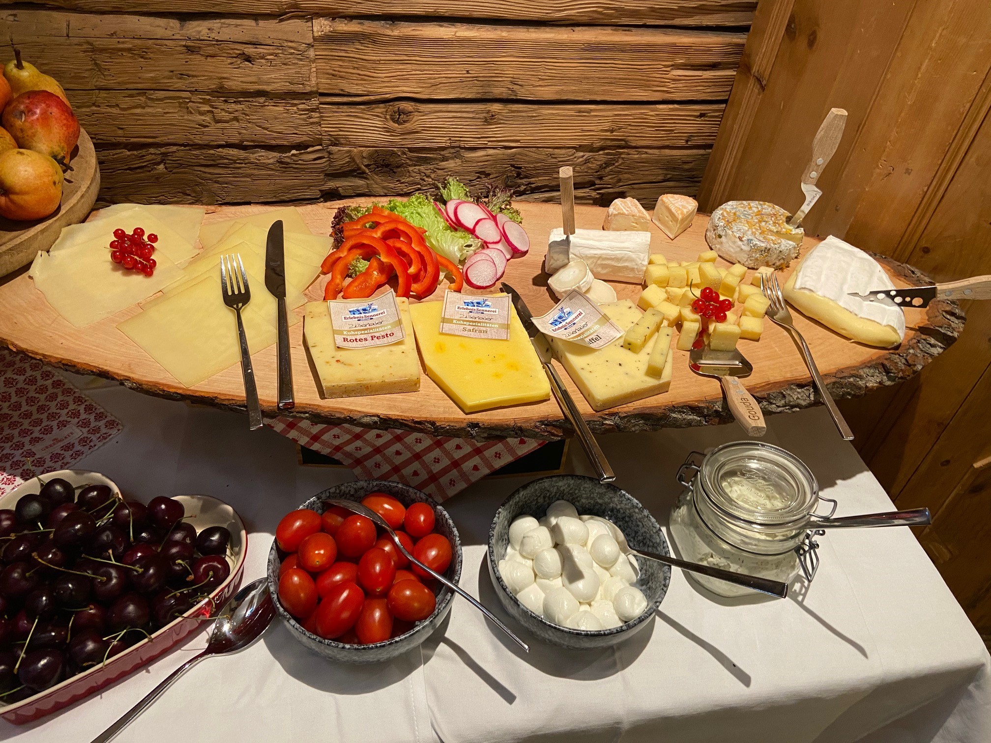 Buffet mit verschiedenen Käsesorten, geschnittenem Gemüse und Brot auf Holzbrett. Davor Schüsseln mit Kirschen, Tomaten und Mozzarella. Rustikales Ambiente.