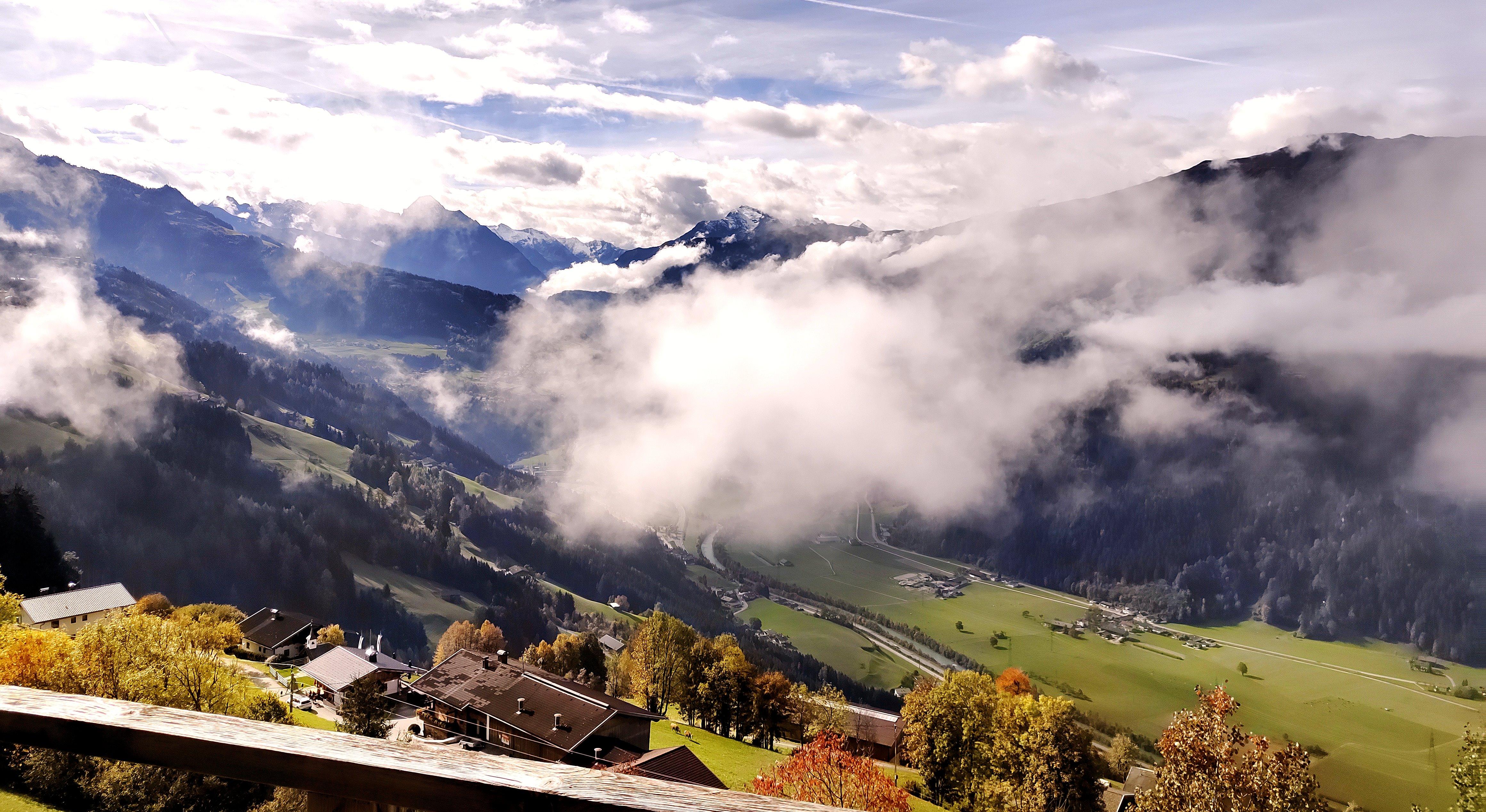 Mountain landscape with clouds, valleys, and houses in the foreground, surrounded by forests and peaks.
