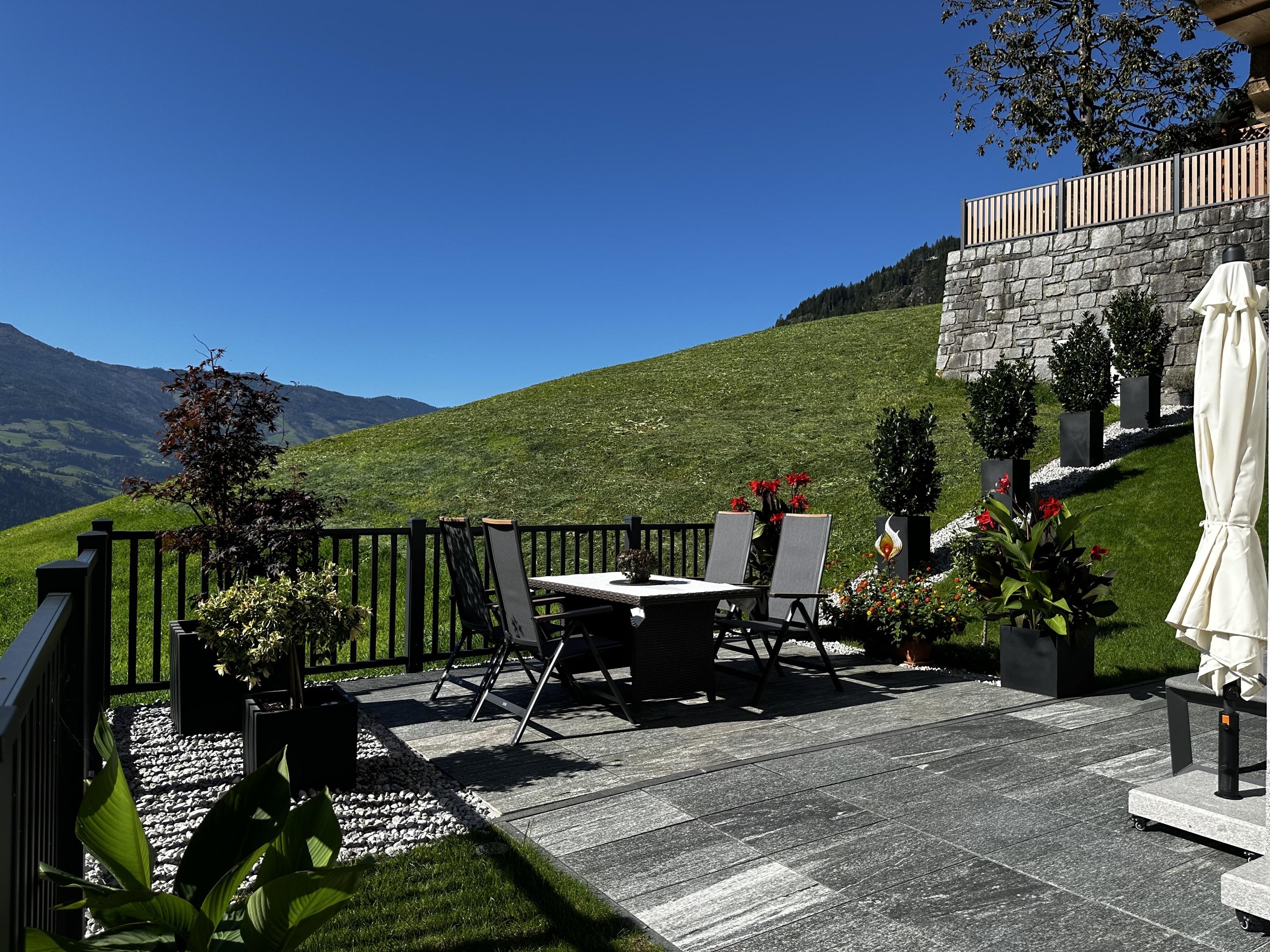 Garden with table, chairs, umbrella, plants, and view of green hills and mountains.