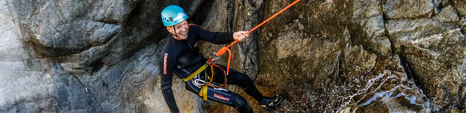 A person wearing a helmet, wetsuit, and harness is abseiling down a rock face while canyoning in the Mayrhofen-Hippach holiday region. They hold a rope and smile as water flows down the rocks.