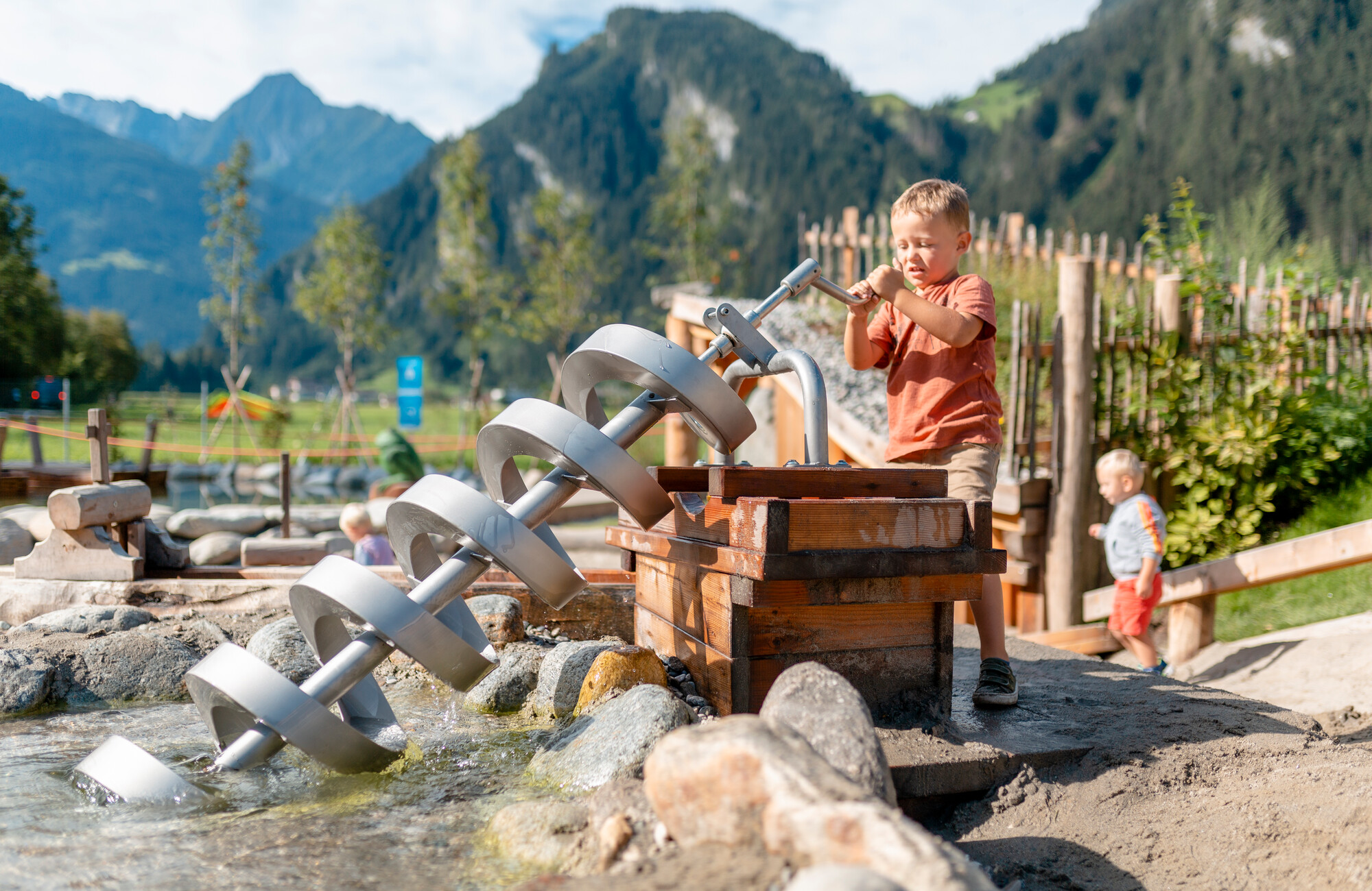 A child enthusiastically plays in the water play area at the Auenland Sidan playground in Schwendau. In the background, another child, wooden play equipment, and the Tyrolean mountain landscape. 