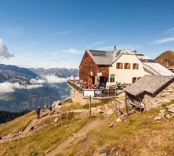 Hütten auf einer Bergkuppe mit Blick auf Wolken und Berge im Hintergrund, bei schönem Wetter.