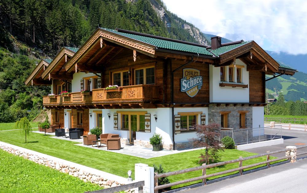 A two-story chalet-style house with wood and white walls, green roof, surrounded by greenery and mountains.
