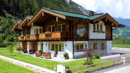 A two-story chalet-style house with wood and white walls, green roof, surrounded by greenery and mountains.