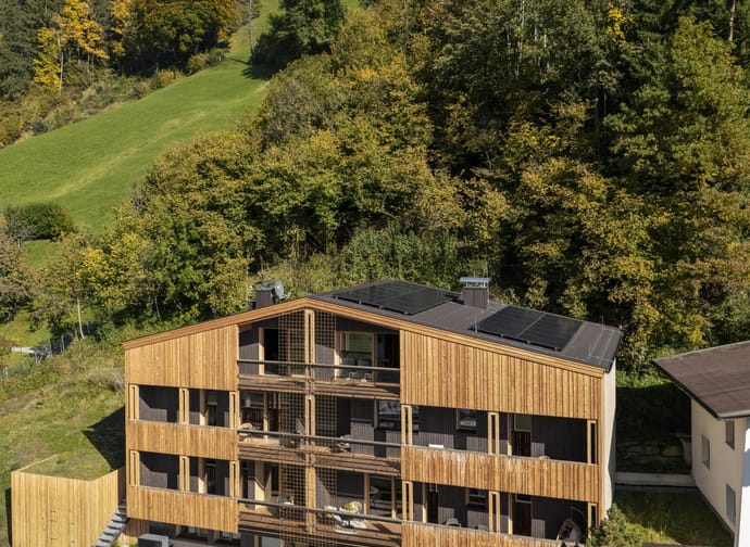 Modern wooden building with balconies in front of a forested hillside and blue sky.