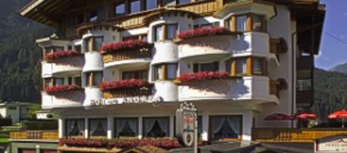 The image shows Hotel Andrea from the outside – a well-kept building in classic Alpine style with wooden balconies and charming details. In the foreground are green plants, with mountains rising up in the background