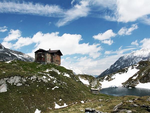 An old house on a green, snow-dusted hillside with mountains and a lake in the background.