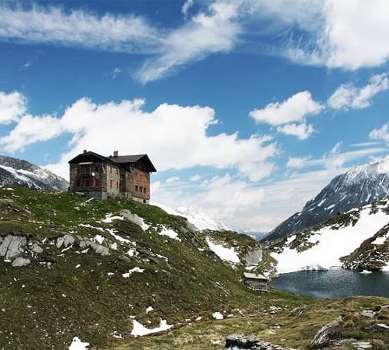 An old house on a green, snow-dusted hillside with mountains and a lake in the background.