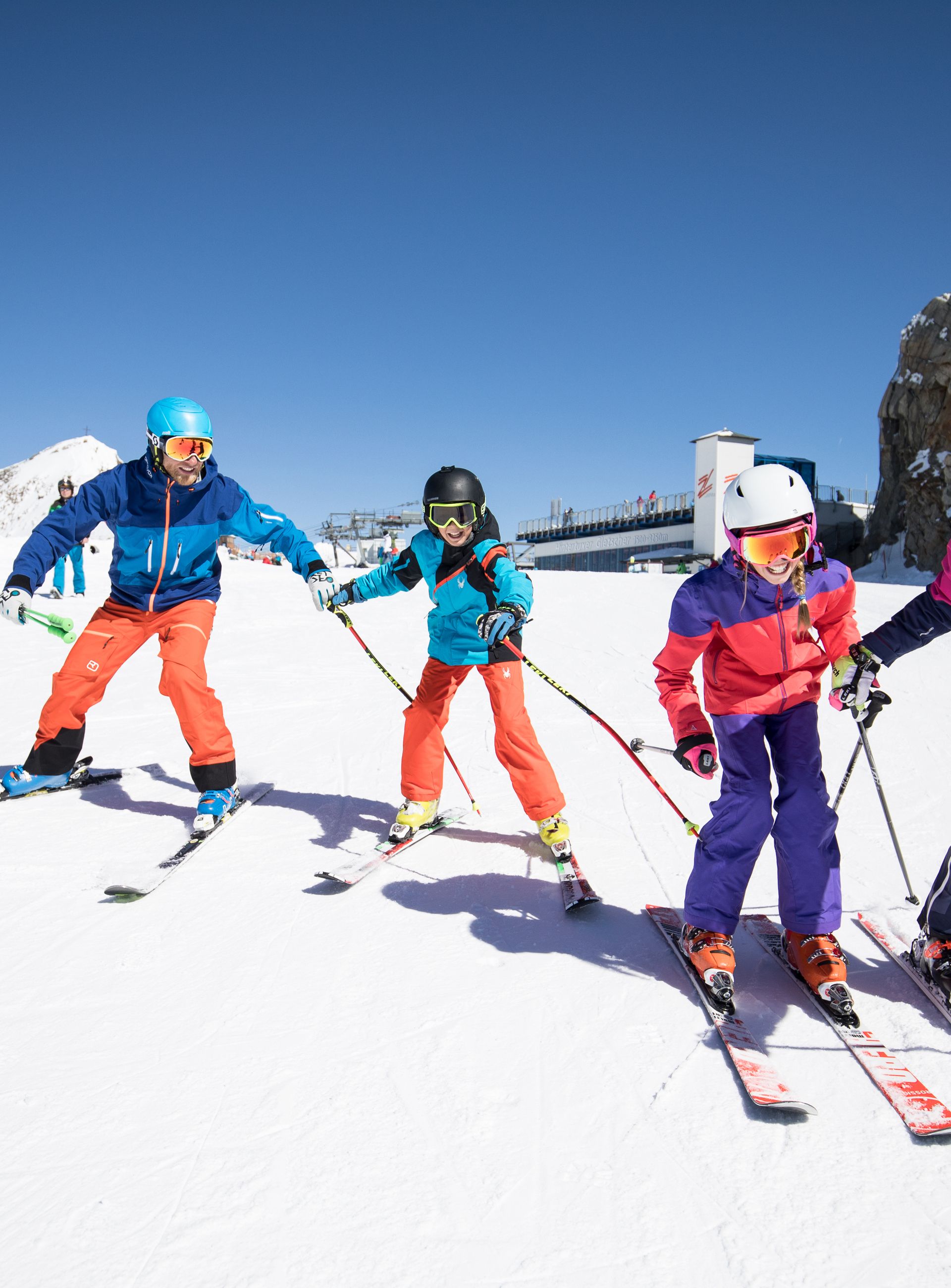 Four skiers in colourful jackets and helmets glide side by side across the snow-covered Hintertuxer Glacier, with bright white snow and a dark rock face behind them under a brilliant blue sky.