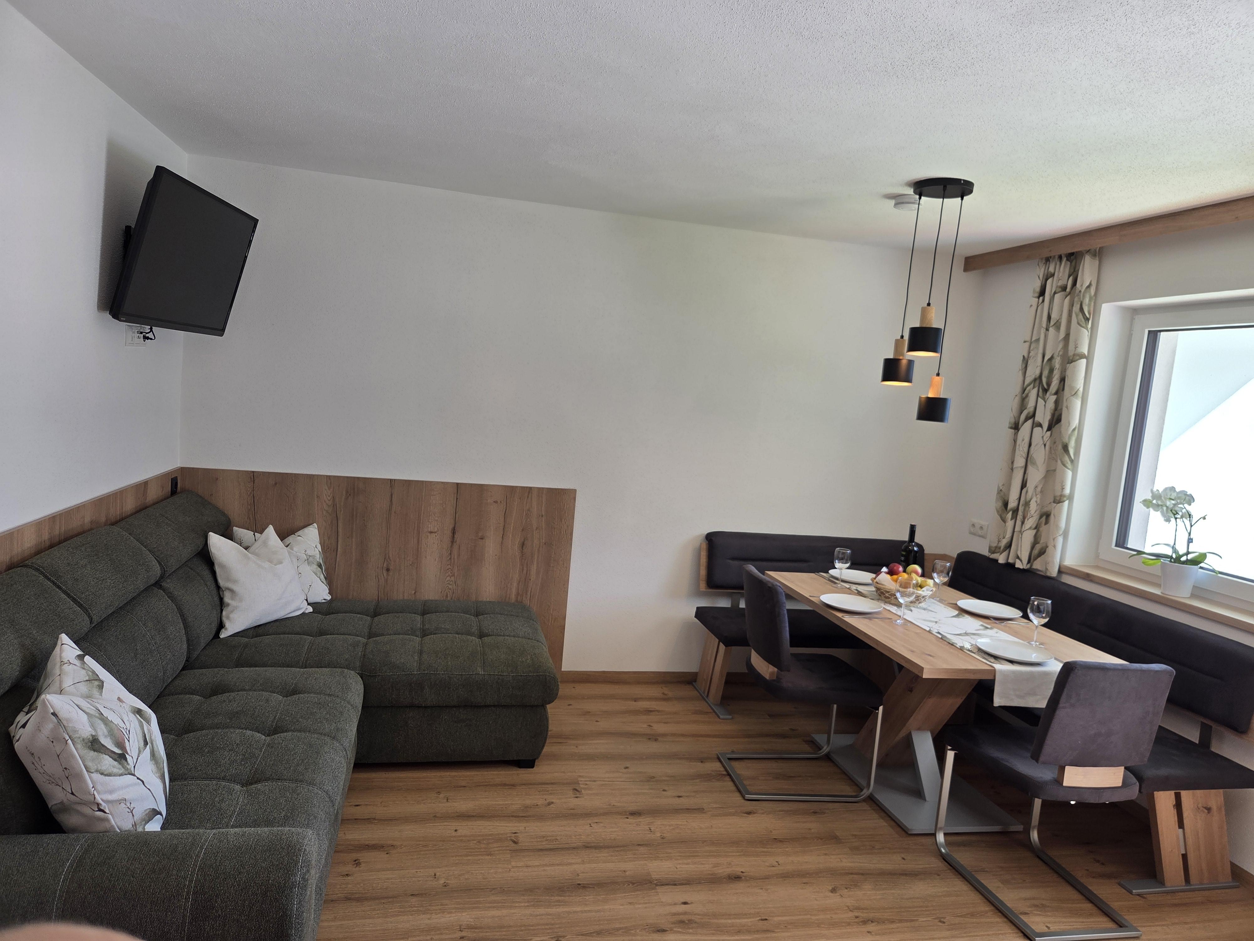 Gemütliches Wohnzimmer mit grauer Couch, Esstisch, Stühlen, Wandlampe und Fenster mit Vorhängen.