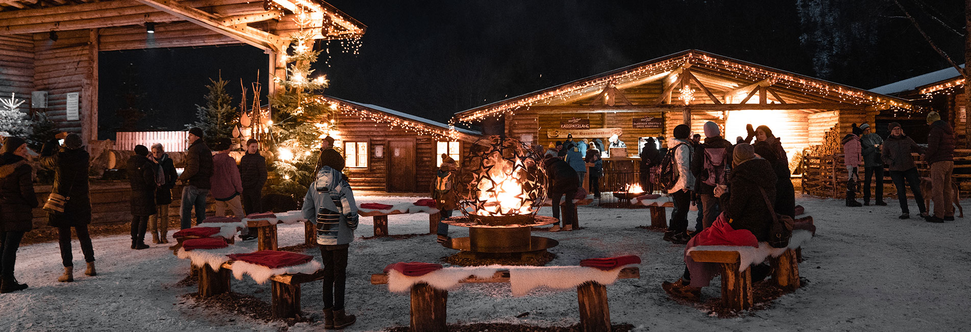 People gather around festive lights and fire outside wooden cabins in a snowy winter scene.