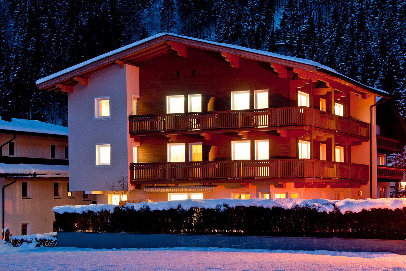 Ein mehrstöckiges Chalet im Schnee mit beleuchteten Fenstern und Holzbalconien in einer Berglandschaft bei Nacht.