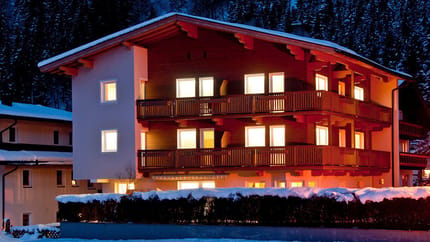Ein mehrstöckiges Chalet im Schnee mit beleuchteten Fenstern und Holzbalconien in einer Berglandschaft bei Nacht.