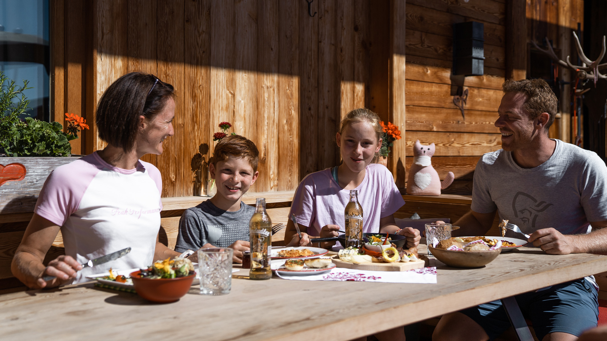 Eine vierköpfige Familie sitzt lachend an einem rustikalen Holztisch auf einer sonnigen Almterrasse im Zillertal vor einer Holzwand und genießt regionale Speisen und Getränke beim Genussherbst.