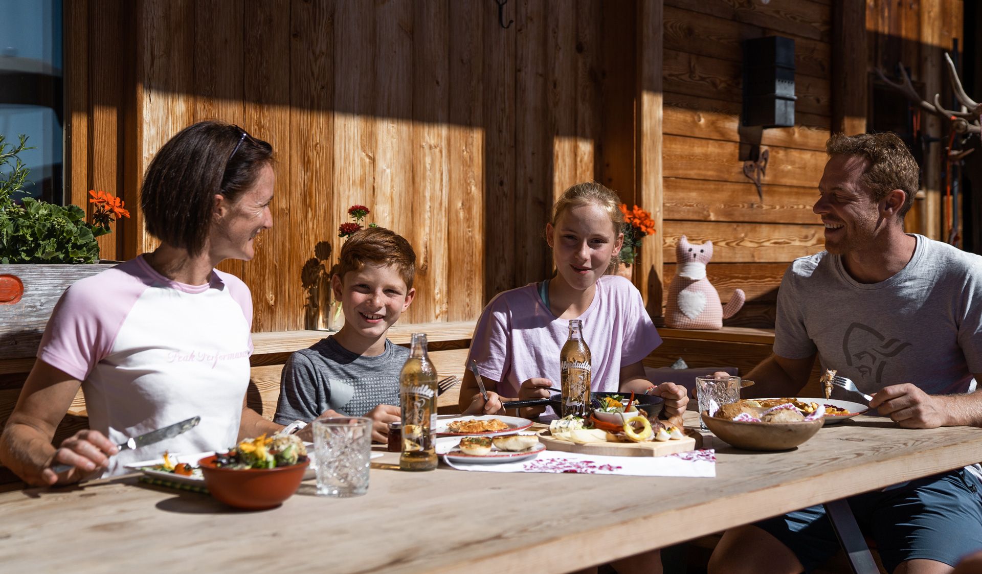 Genuss und Kulinarik in Mayrhofen-Hippach Familie sitzt draußen an Holztisch vor Berghütte und isst gemeinsam. Sonniges Wetter, Berge und Natur im Hintergrund, freundliche Atmosphäre.
