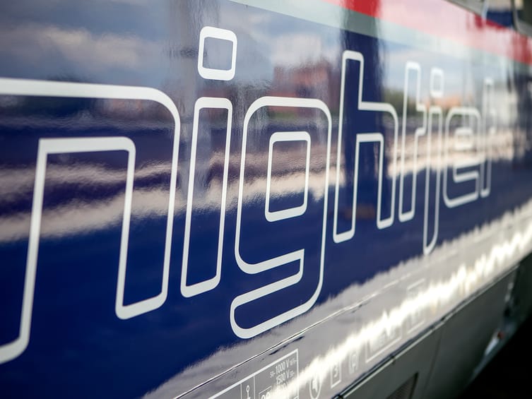 Nahaufnahme der weißen 'Nightjet' Beschriftung auf der blauen Seitenwand eines ÖBB Nachtzuges. Die moderne Typografie ist deutlich sichtbar auf der glatten Zugoberfläche.