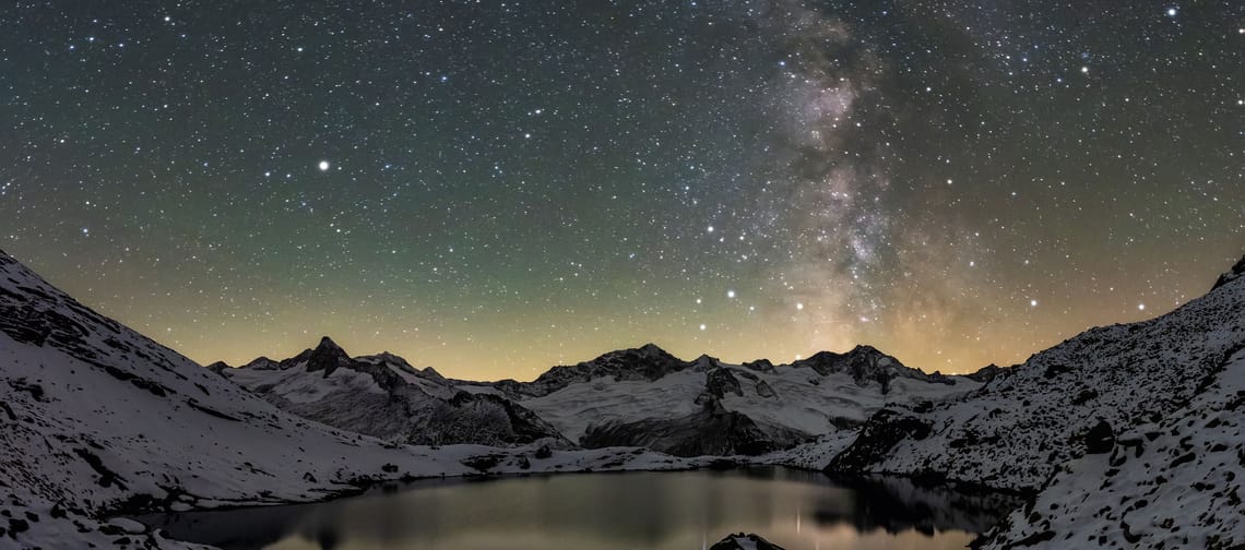 Starry night sky over a mountain lake with snow-capped peaks in the foreground.