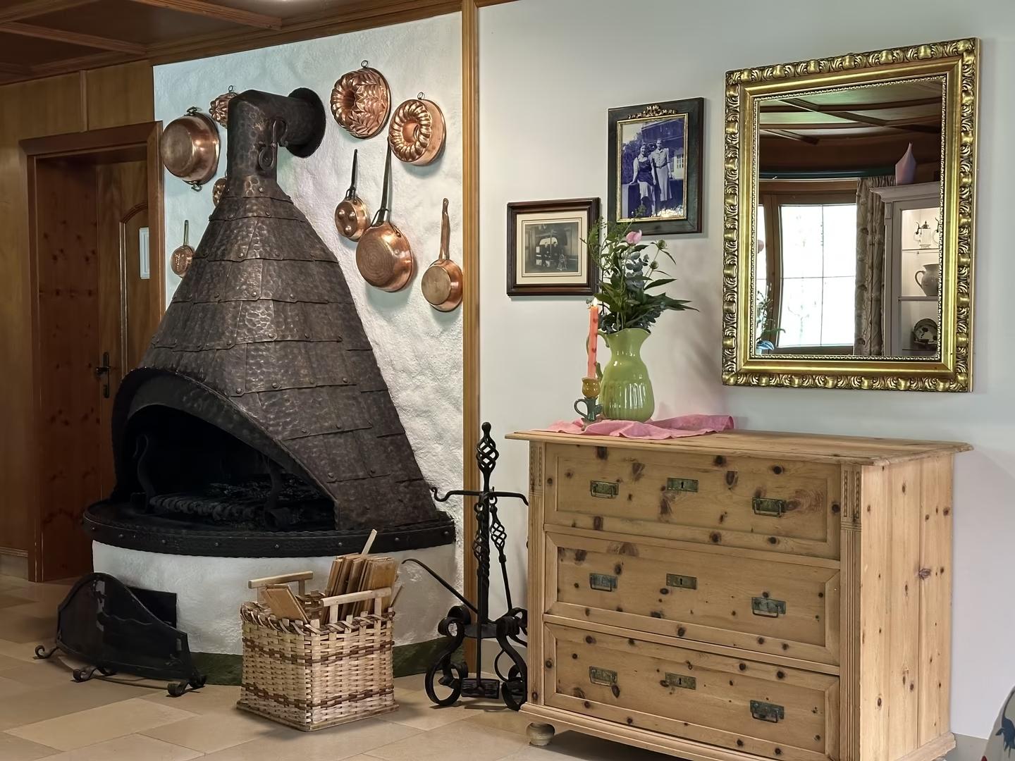 Rustikaler Kamin mit Kupfertöpfen an der Wand, Holzkommode, Spiegel und Vase mit Blumen im Raum.