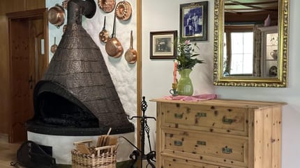Rustikaler Kamin mit Kupfertöpfen an der Wand, Holzkommode, Spiegel und Vase mit Blumen im Raum.