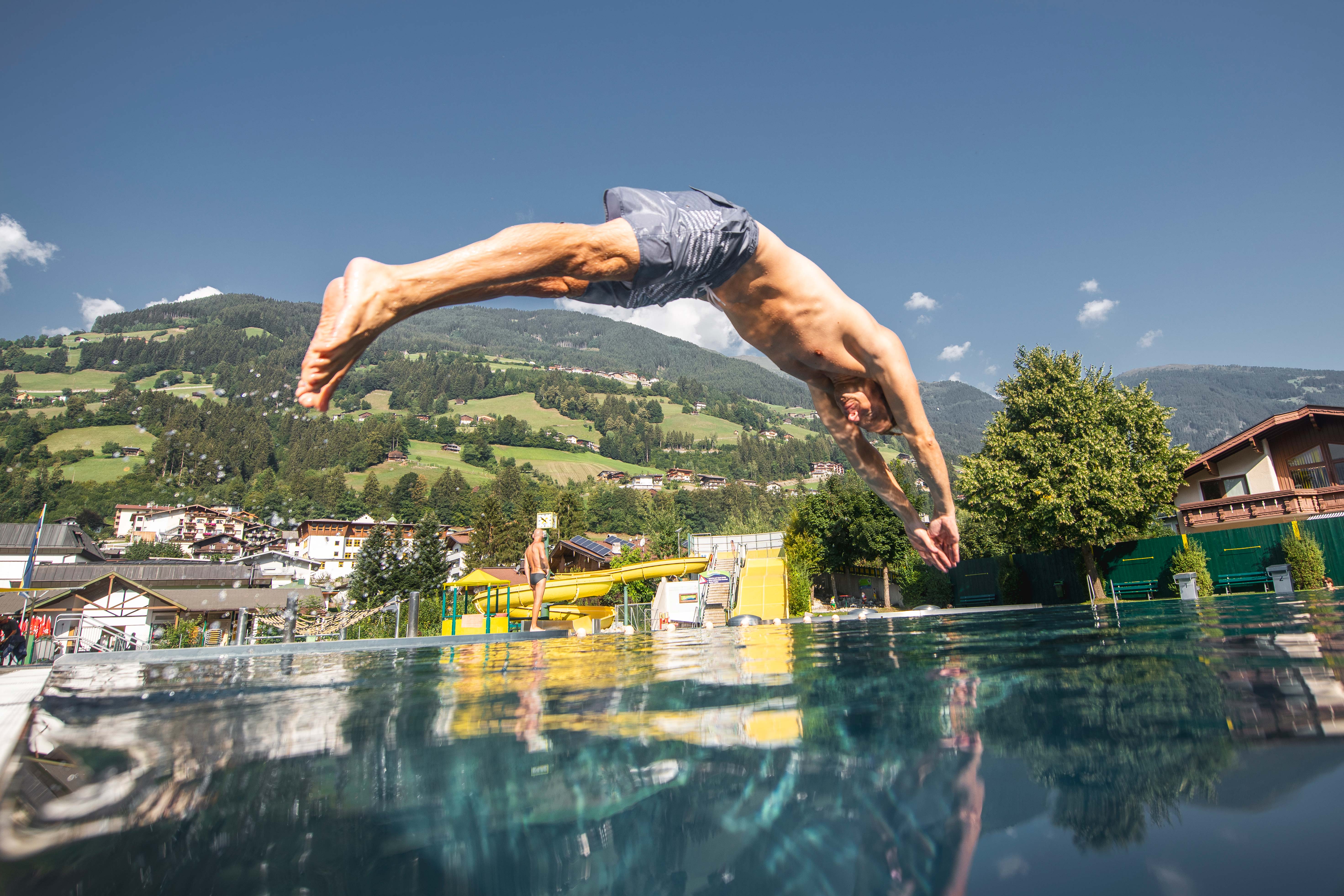 Freischwimmbad Hippach – Mann springt ins Wasser vor alpiner Kulisse bei strahlendem Sonnenschein. Sommerlicher Badespaß, Bewegung und Natur im Zillertal, Tirol.