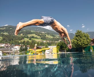 Mann springt mit gestrecktem Körper ins Wasser des Freibads Hippach an einem sonnigen Tag, mit Blick auf Berge und grüne Landschaft.
