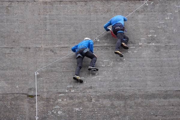 Zwei Männer mit blauer Jacke und schwarzer Hose klettern entlang der Staumauer auf den Eisenhaken.