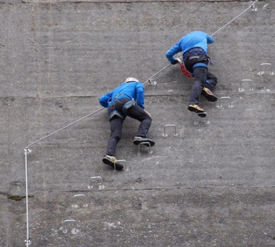Zwei Männer mit blauer Jacke und schwarzer Hose klettern entlang der Staumauer auf den Eisenhaken.