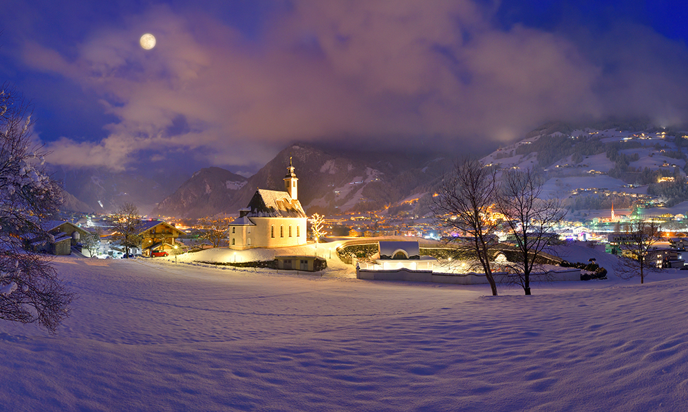 Nächtliche Ortsansicht von Ramsau im Zillertal. Eine hell erleuchtete Kirche steht in tief verschneiten Feldern. Im Hintergrund funkeln die Lichter des Dorfes vor dunklen Bergen unter einem hellen Vollmond.