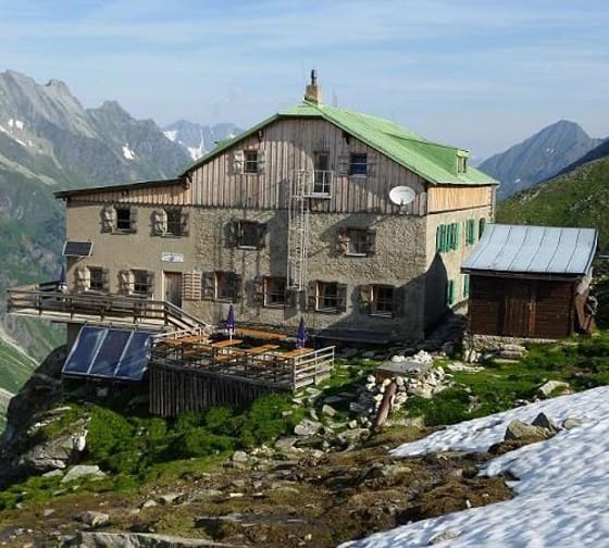 Mountain hut with wooden and stone exterior, surrounded by snow and mountains, with solar panels and satellite dish.