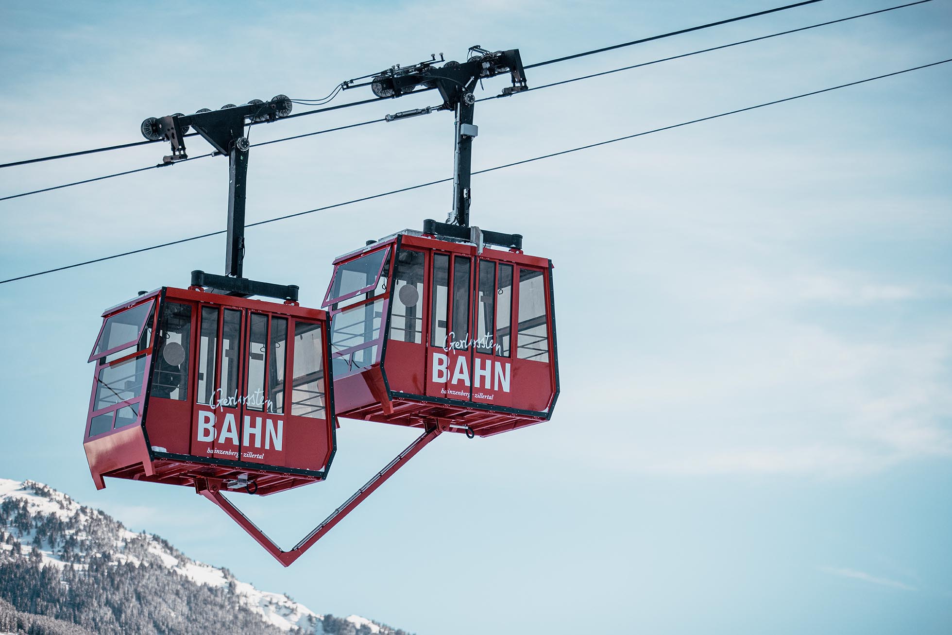 Zwei rote Gondeln der Seilbahn vor einem Berg mit Schneeflächen, Himmel im Hintergrund.