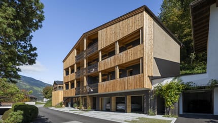 Modern wooden and concrete building with balconies, surrounded by trees and mountains under a clear sky.