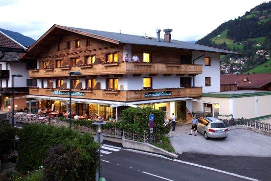 Das Bild zeigt den Gasthof Zillertaler Weinstadl von außen. Das Gebäude besteht überwiegend aus Holz und strahlt eine gemütliche Atmosphäre aus. Vor dem Gasthof steht ein Auto auf dem Parkplatz, im Hintergrund sind Berge unter einem grauen Himmel zu sehen