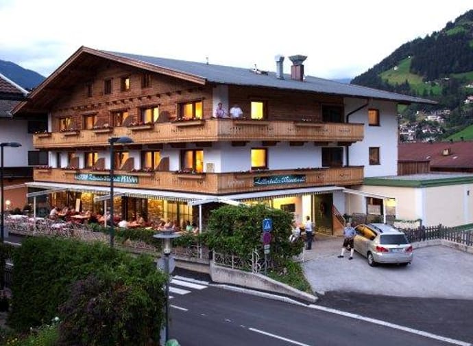 The picture shows Gasthof Zillertaler Weinstadl from the outside. The building is mainly made of wood, radiating a cozy atmosphere. A car is parked in front of the inn, and in the background, mountains are visible under a gray sky.