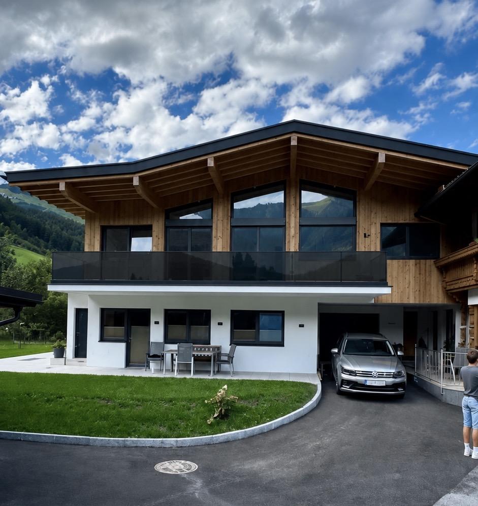 Modernes zweistöckiges Haus mit Holz und Weiß, Balkon, Auto im Carport, grüner Garten, bei bewölktem Himmel.