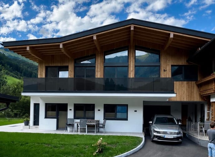 Modernes zweistöckiges Haus mit Holz und Weiß, Balkon, Auto im Carport, grüner Garten, bei bewölktem Himmel.