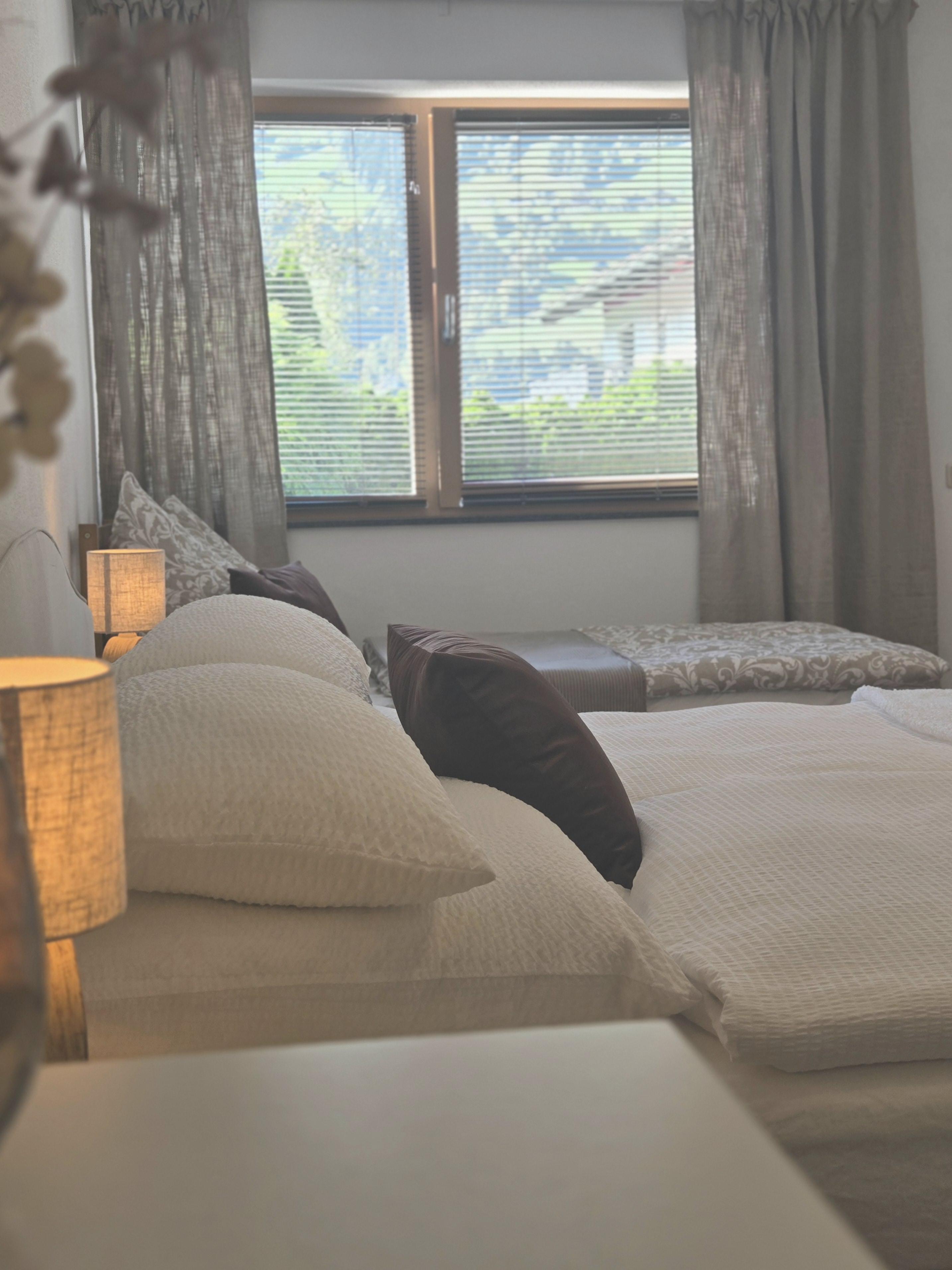 Bett im Schlafzimmer mit weißen Bettdecken, dunklen Kissen und einem Fenster mit Vorhängen im Hintergrund.