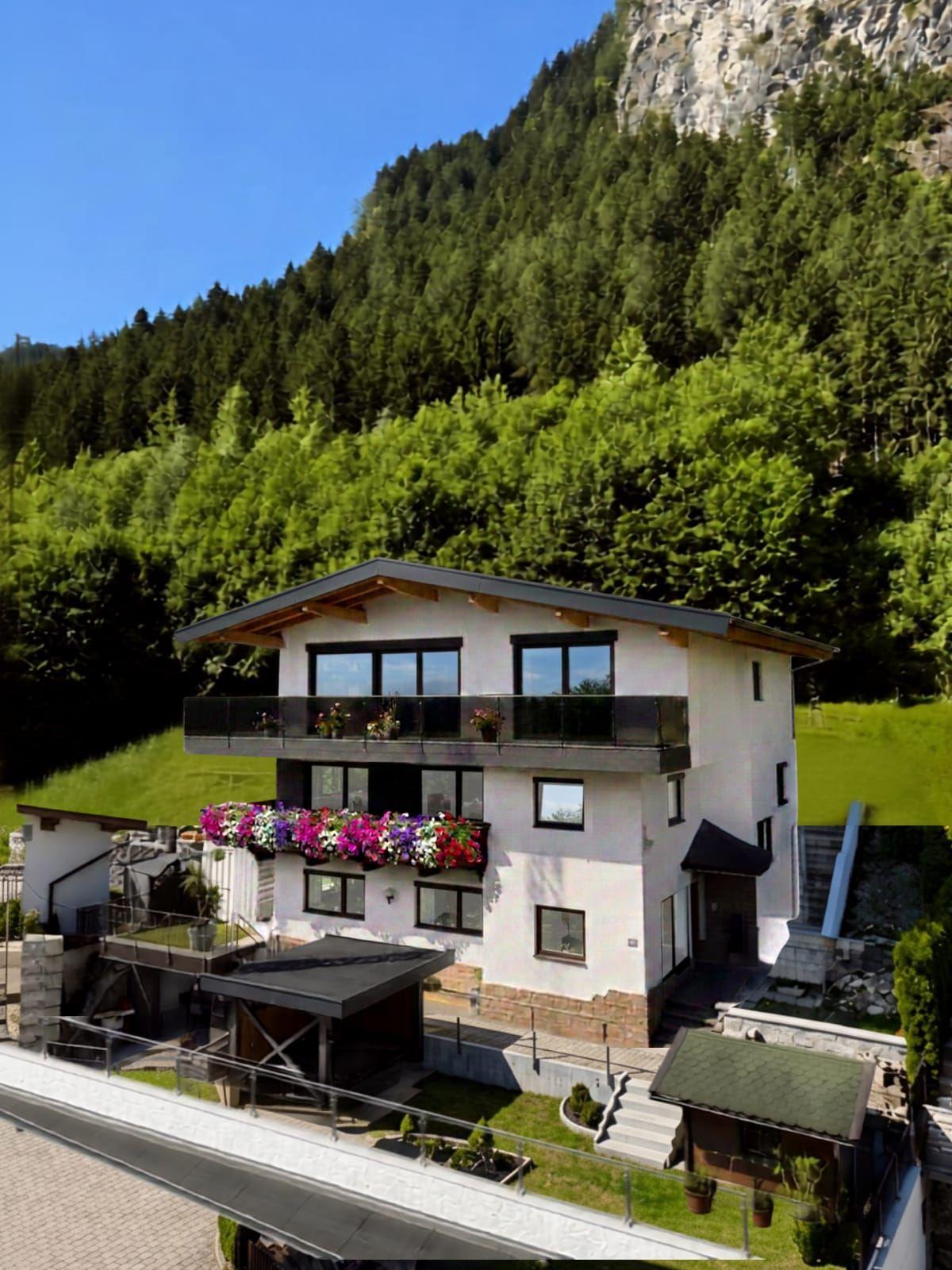 Modernes Haus mit Balkon und Blumen vor grüner, bewaldeter Bergkulisse bei blauem Himmel.
