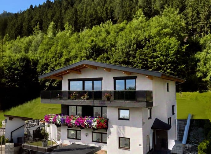 Modernes Haus mit Balkon und Blumen vor grüner, bewaldeter Bergkulisse bei blauem Himmel.